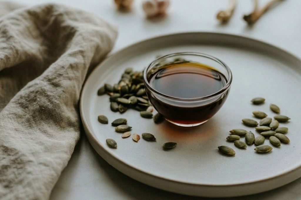 Small glass dish of dark Styrian pumpkin seed oil beside dark green seeds on pale ceramic